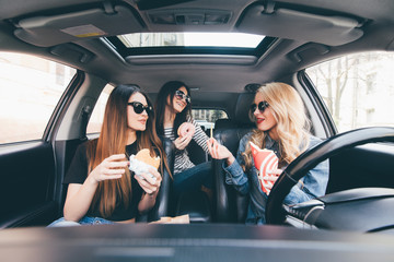 Three young women drive a car, speaking each other and eating a fast food in a car in traffic jam