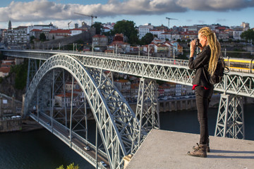Traveller girl photographer with blonde dreadlocks is takes photo Dom Luis I bridge over Douro river, Porto, Portugal.