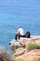 Mann in Jeans und T-Shirt mit Rucksack einsam auf der Klippe sitzend, den Blick auf das Meer gerichtet