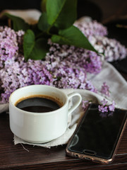 A cup of coffee on the table and lilac flowers and a mobile phone