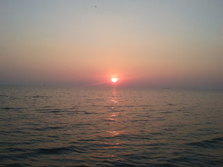 Obraz premium A blurred disk of bright sun over horizon against the background of a reddish sky. Sea sunset with reflection at water.