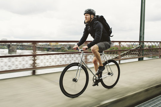 Male Commuter Riding Bicycle On Bridge Over River Against Cloudy Sky