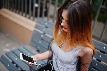Fototapeta na wymiar Fashionable woman look at white shirt, black transparent clothes, leather pants, posing at street and sitting on bench with cell phone and headphones.