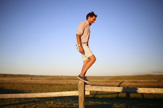 Full length of happy man balancing on wooden fence against clear blue sky during sunset
