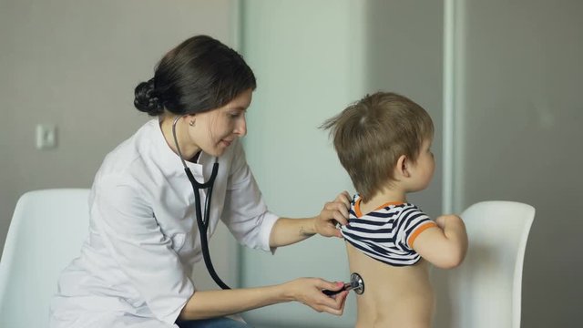 Young woman doctor talking little boy and listening with stethoscope in medical office