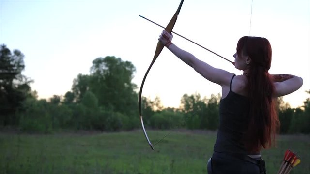 Female ginger hair archer shooting targets with her bow and arrow with self made mehendi henna tatoo on hand. Concentration concept.