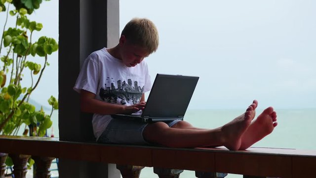 young guy playing on a laptop, sitting on the terrace. Near the sea and the beach