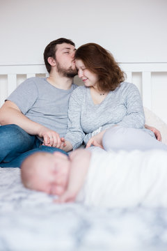 Young parents resting on white bed, kissing, while their newborn girl lying in the foreground. Natural, lifestyle image with shallow depth of field.