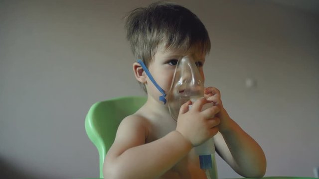 Sick child breathes through nebulizer inhalation, boy with an oxygen mask on his face having treatment at home