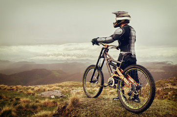 The rider in the full-face helmet and full protective equipment on the mtb bike stands on a rock against the background of a ridge and low clouds