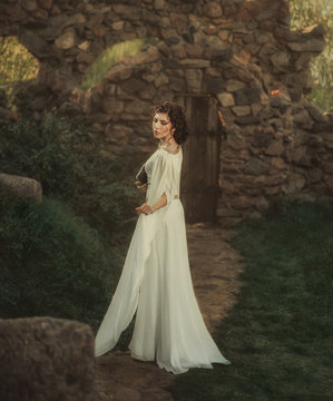 Young vintage woman walk near castle. adult beautiful girl in Greek white medieval historical dress holding book in hand.  princess stands, old stone courtyard path way.  warm colors back rear view