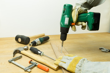Hands in brown and gray work gloves using a green power battery drill screwdriver on a working bench with tools spread on it