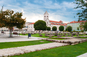 Fototapeta na wymiar Tower of St. Mary's church, the forum square and stone ruins among green lawn and trimmed trees on a summer morning. Dramatic cloudy sky. Solitary and cozy Zadar center, Croatia