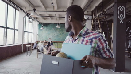 Young african man get fired from work at start-up. Male walks through the office, carrying box with...