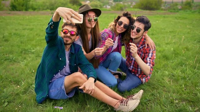 Portrait of four happy hipsters while they making selfie with sparklers on a smartphone. Cheerful people. Summer lifestyle
