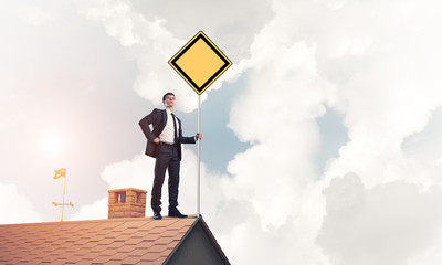 Young businessman on house brick roof holding yellow signboard. 