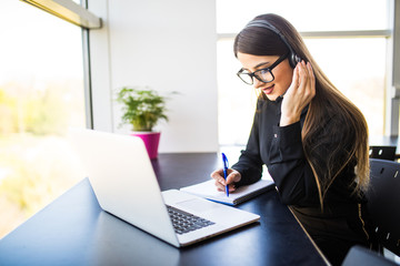 thrilled young woman manager taking notes on good news on headset and notebook in office