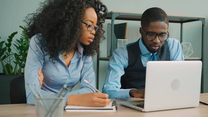 Two african american business people working together in in the office. African American businessman and businesswoman talking, using laptop and writing down information. Business team - Powered by Adobe