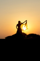 A girl in Indian clothes dances at sunset in the desert on the dunes against low bushes, sun and sky. Rajasthan, India.