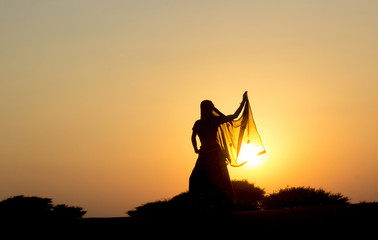Young woman  in Indian clothes dances at sunset in the desert on the dunes against low bushes, sun and sky. Rajasthan, India.