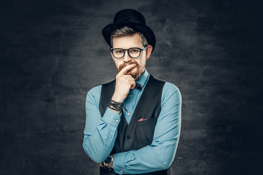 An elegant bearded hipster male dressed in a blue shirt, waistcoat and cylinder hat over grey vignette background.