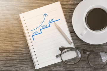 arrow pointing up with Yes You Can - Motivation Business Concept  message on notebook with glasses, pencil and coffee cup on wooden table.