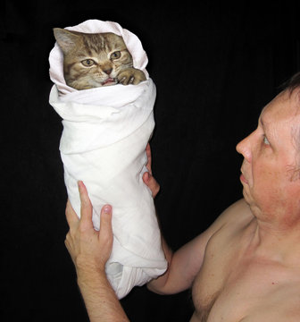 The man is holding a kitten. It is in swaddling clothes. Black background.