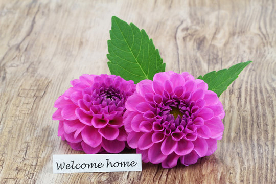 Welcome home card with two pink dahlia flowers on wooden surface
