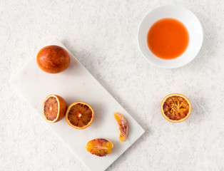Whole, halved and squeezed blood oranges and fresh juice on white marble chopping board and white textured marbled background with copy space.