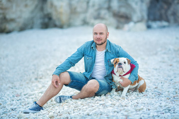 Handsome bold stylish dressed in jeans man in blue cap sitting on a beach sea side together with pretty english bulldog.
