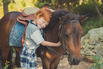 Cute red hair boy smiling to camera and standing in forest with horse