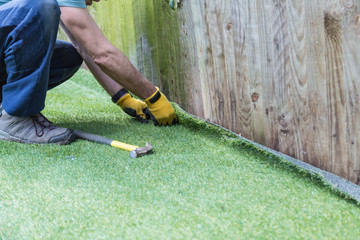 Artificial grass being installed. It has been cut to size and rolled out and laid and is being nailed down along a fence. The installer has a hammer and nails. - 153825143