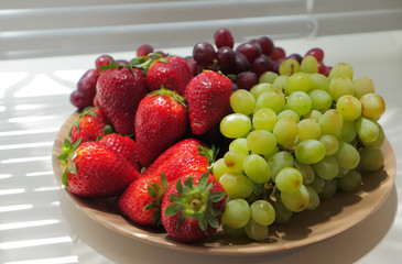 Close up of fresh red strawberry in bowl with green and red grapes behind the window.  Fresh strawberries on the plate beside the window in the morning. 