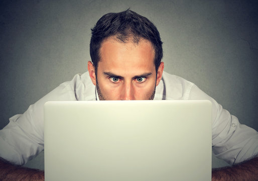 Man hiding behind a laptop staring at screen with a shocked face expression