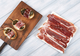 Top view of Spanish tapas of ham and green pepper, and a plate of Iberian ham on white wooden board. Spanish gastronomy.