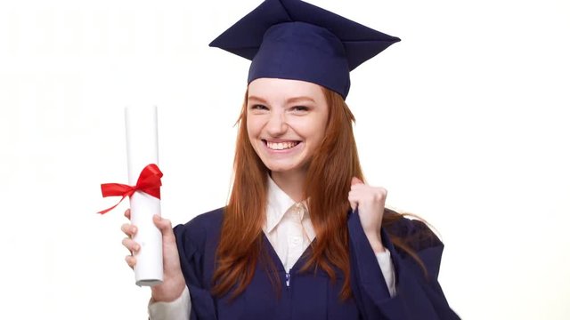 Young happy redhead Cacasian graduate girl happily showing her diploma on white background wearing blue robe and square academical cap