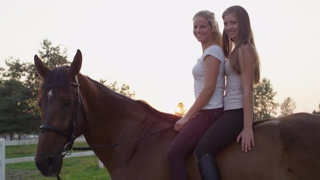 SLOW MOTION, CLOSE UP: Two cheerful young girls sitting one after another on strong gelding in park at sunrise. Girlfriends bareback riding stunning stallion on horse ranch at gorgeous summer sunset