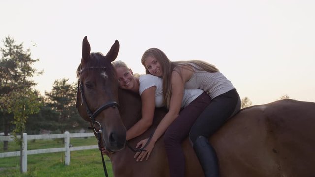 SLOW MOTION, CLOSE UP: Two cheerful young girls sitting on strong chestnut gelding and embracing it. Pretty girlfriends bareback riding stunning brown stallion on horse ranch at gorgeous summer sunset