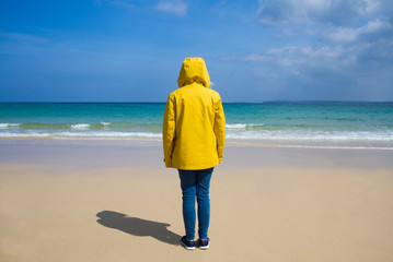 A rear view of a lone woman wearing a bright yellow hooded jacket and facing away from the camera towards the ocean on a deserted, sandy beach.