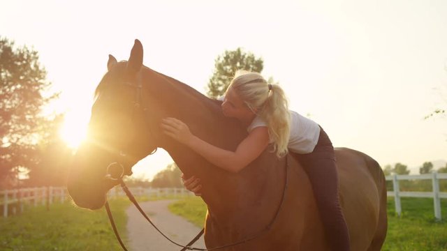 SLOW MOTION, CLOSE UP, DOF: Pretty blonde girl horseback riding beautiful dark brown gelding, embracing and caressing him. Happy young woman bareback riding powerful stallion at gorgeous golden sunset