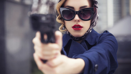 Beautiful brunette sexy spy agent (killer or police) woman in jacket and sunglasses with a gun in her hand runningr someone, to catch him in a european city