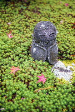Stone statue of Jizo on ground covered by green star moss and red maple leaves during autumn in a garden at Enkoji temple in Kyoto, Japan