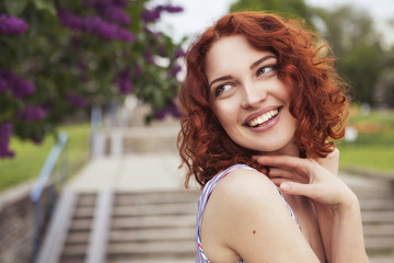 Beautiful red haired woman with fresh flawless skin and curly hairstyle in dress and a hat, smiling cheerfully in park. ideal teeth. Sunny day