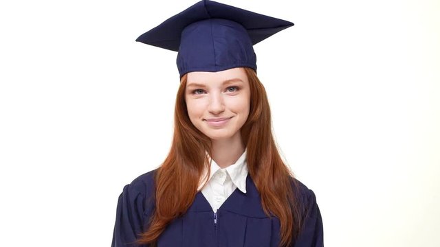 Very cute foxy Caucasian graduate girl in blue robe and square academical cap quietle standing on white background in slowmotion and smiling