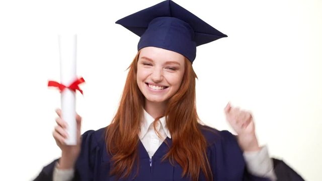 Young happy redhead Cacasian graduate girl happily showing her diploma on white background wearing blue robe and square academical cap in slowmotion