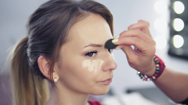 Closeup of professional makeup artist applying concealer to skin of young woman with brush to cover dark circles and make face look brighter. Slowmotion shot