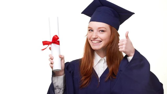 Smiling ginger young graduate girl in blue robe and square academical cap entering frame holding her diploma. Footage on white background in slowmotion