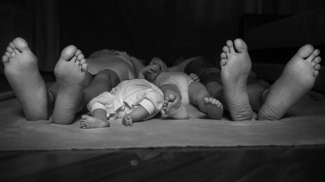 Happy family laying on the floor, their feets on focus. Mother, father, 2 years old daughter and infant newborn baby son. Neutral black background, black and white picture. All in white clothes