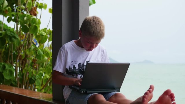 young guy playing on a laptop, sitting on the terrace. Near the sea and the beach