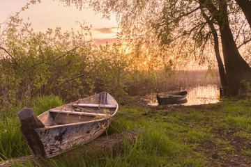 A misty summer morning on the river or lake. Old rough wooden fishing boats on green grass and water, sunrise, fog. Lonely calm mood nature concept. Copy space, horizontal wallpaper or background.